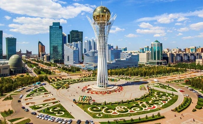 Aerial view of Astana's Bayterek Monument with surrounding plaza, modern skyscrapers, and landscaped gardens under blue sky